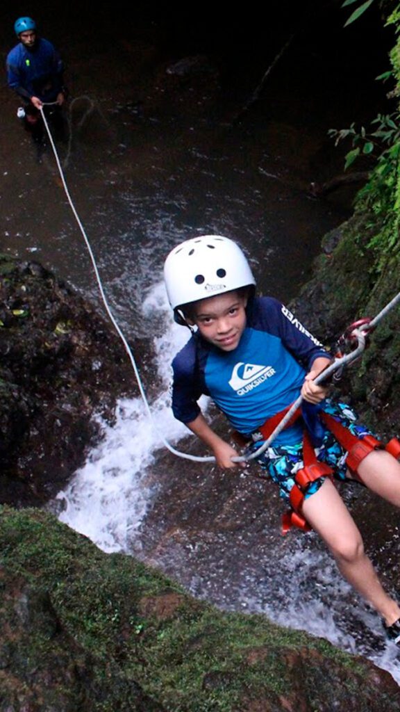 Boy practicing canyoning in Costa Rica rainforest, wearing helmet and harness while descending a waterfall — family adventure activity for kids.